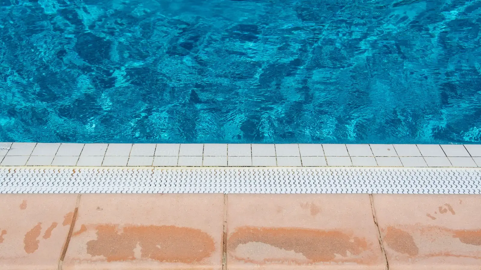 Pool edge from above: rippled blue water, a row of white waterline tiles, an overflow grate and stone coping