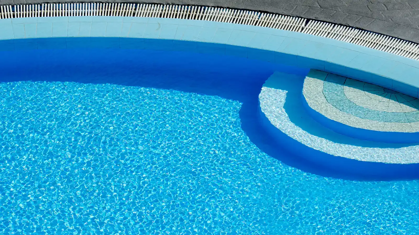 A curved freeform pool from above, with rounded entry steps in one corner and an overflow grate along the edge